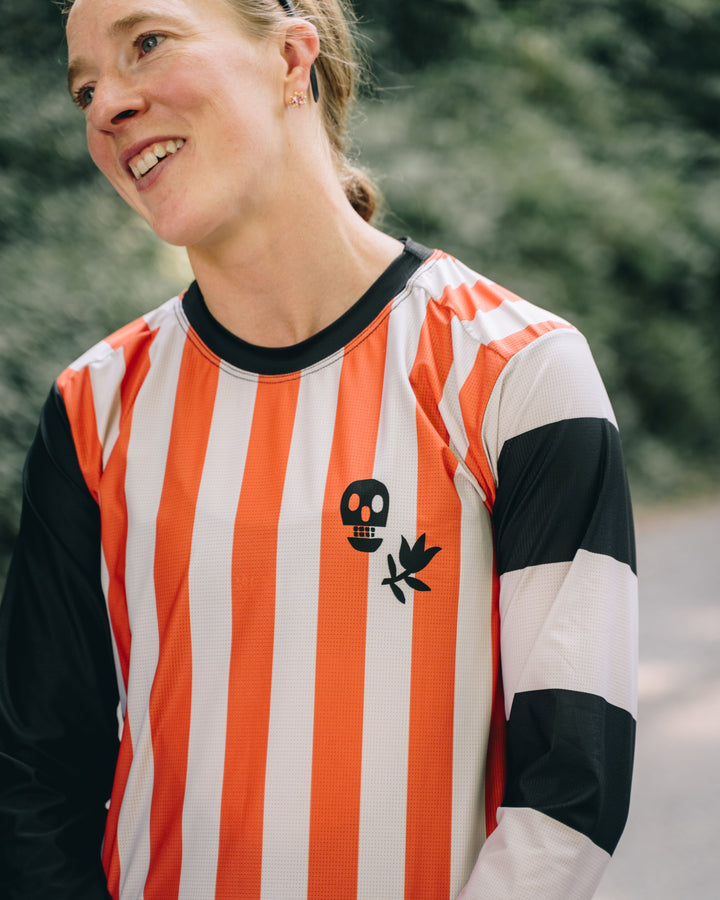 The Women's Harlequin Tech Tee is a long-sleeve, moisture-wicking shirt designed for colder runs or gravel rides. Featuring a bold harlequin stripe pattern, this limited-run tee pairs well with black Death Palm socks and The Athletic cycling bibs. Crafted in California from Italian fabrics by Endo Customs, it offers both style and performance for active individuals