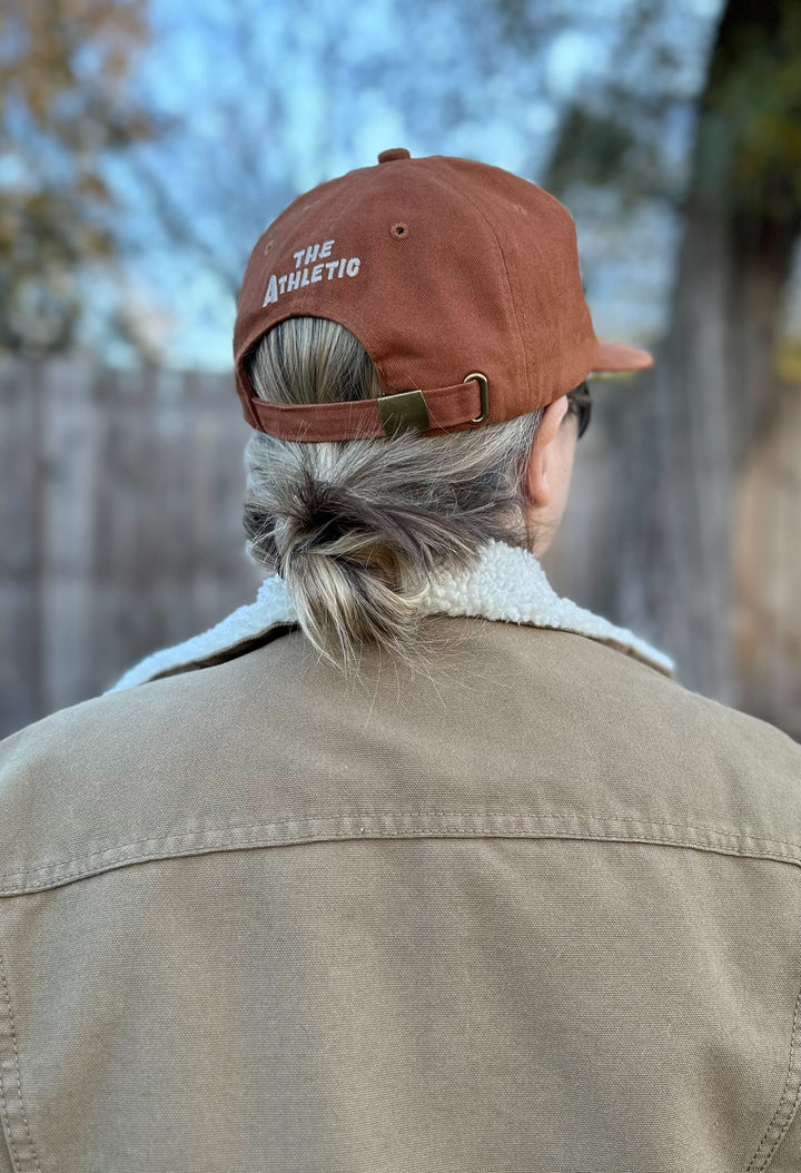 The Athletic Armadillo Logo Hat in rust features an unstructured 6-panel design with an adjustable fabric strap and brass metal clasp. Embroidered with a stylized armadillo logo, this hat combines comfort and style for everyday wear