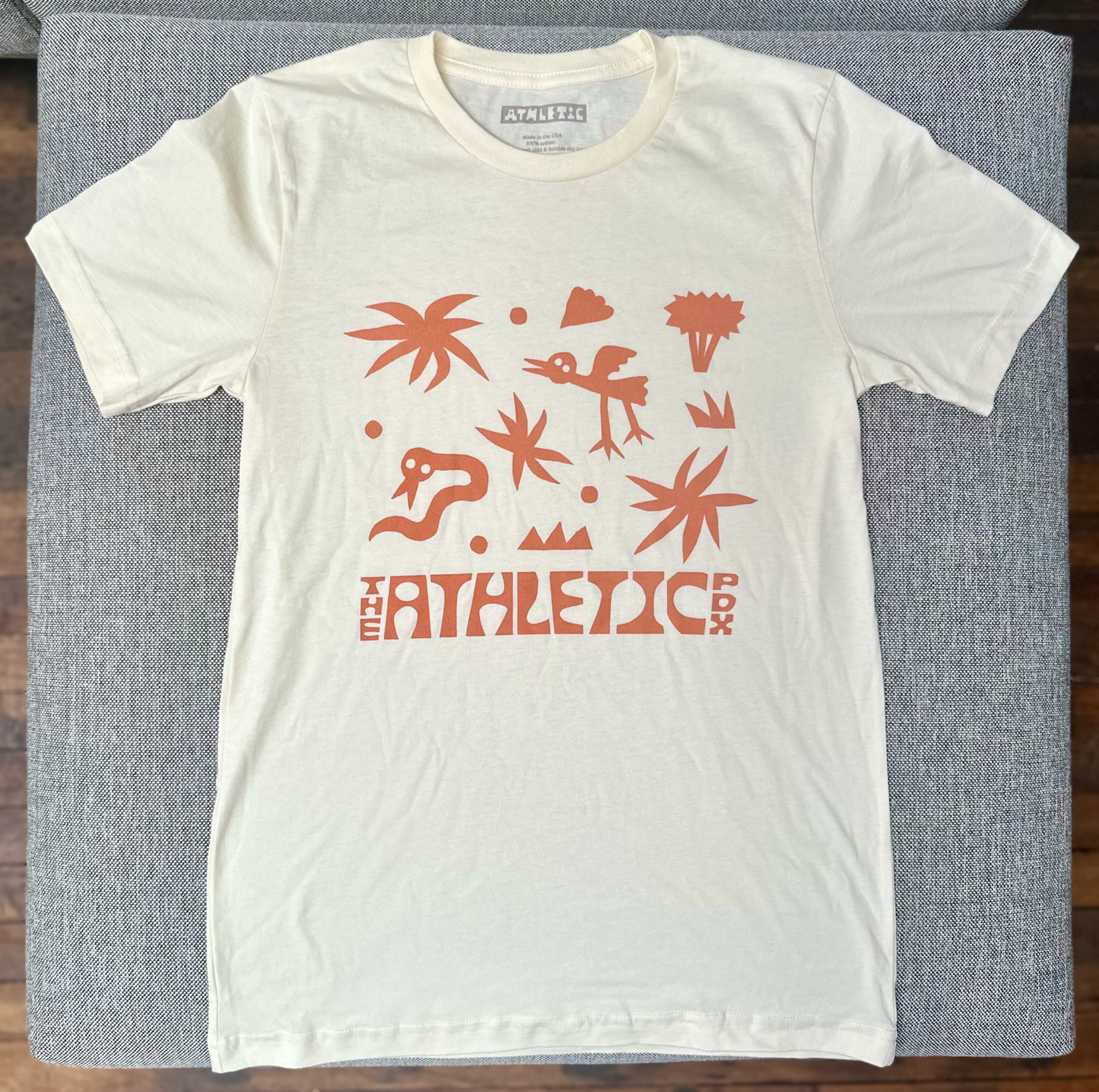 natural color t-shirt laid flat with jungle pattern design with snake and bird and flora that says "the athletic pdx"