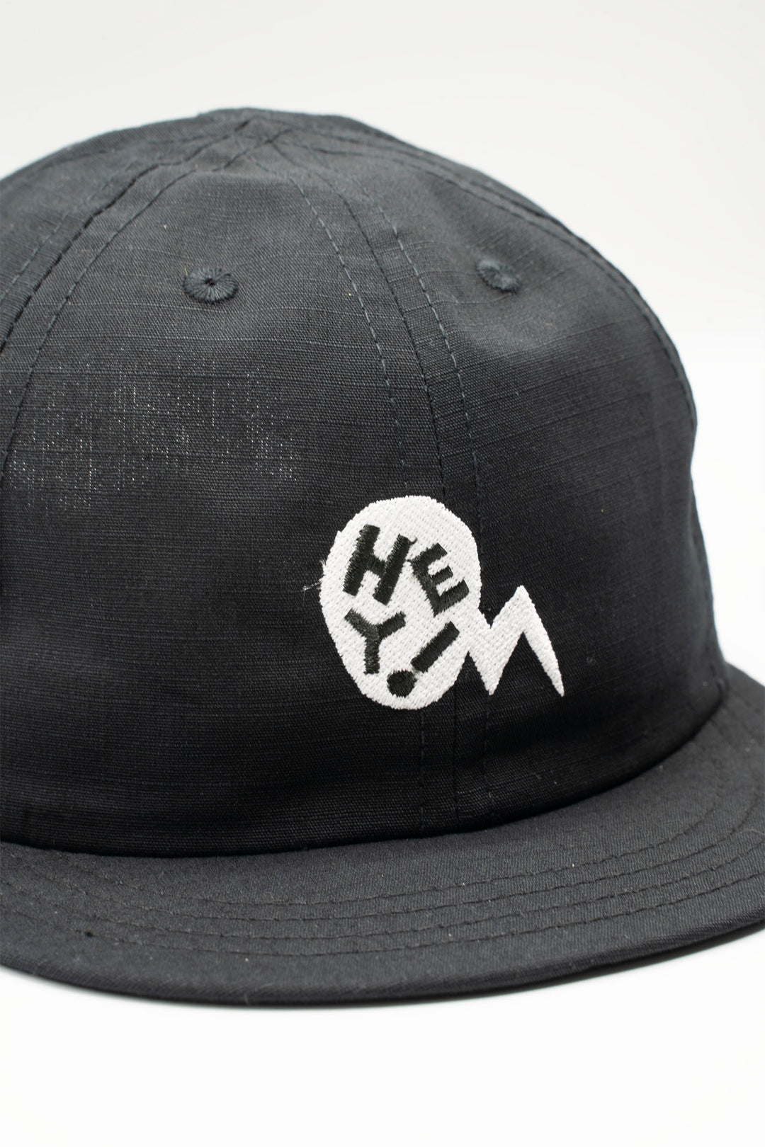 The Hey! Cap is a short-billed, packable hat designed for cyclists and outdoor enthusiasts. It features a black fabric with a white embroidered "Hey!" bubble, offering a stylish and functional accessory for on and off the bike. Proudly made in the USA