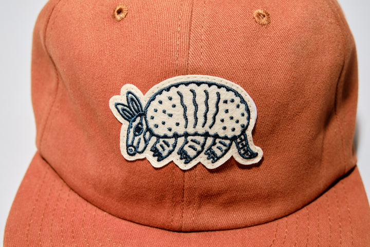 The Athletic Armadillo Logo Hat in rust features an unstructured 6-panel design with an adjustable fabric strap and brass metal clasp. Embroidered with a stylized armadillo logo, this hat combines comfort and style for everyday wear