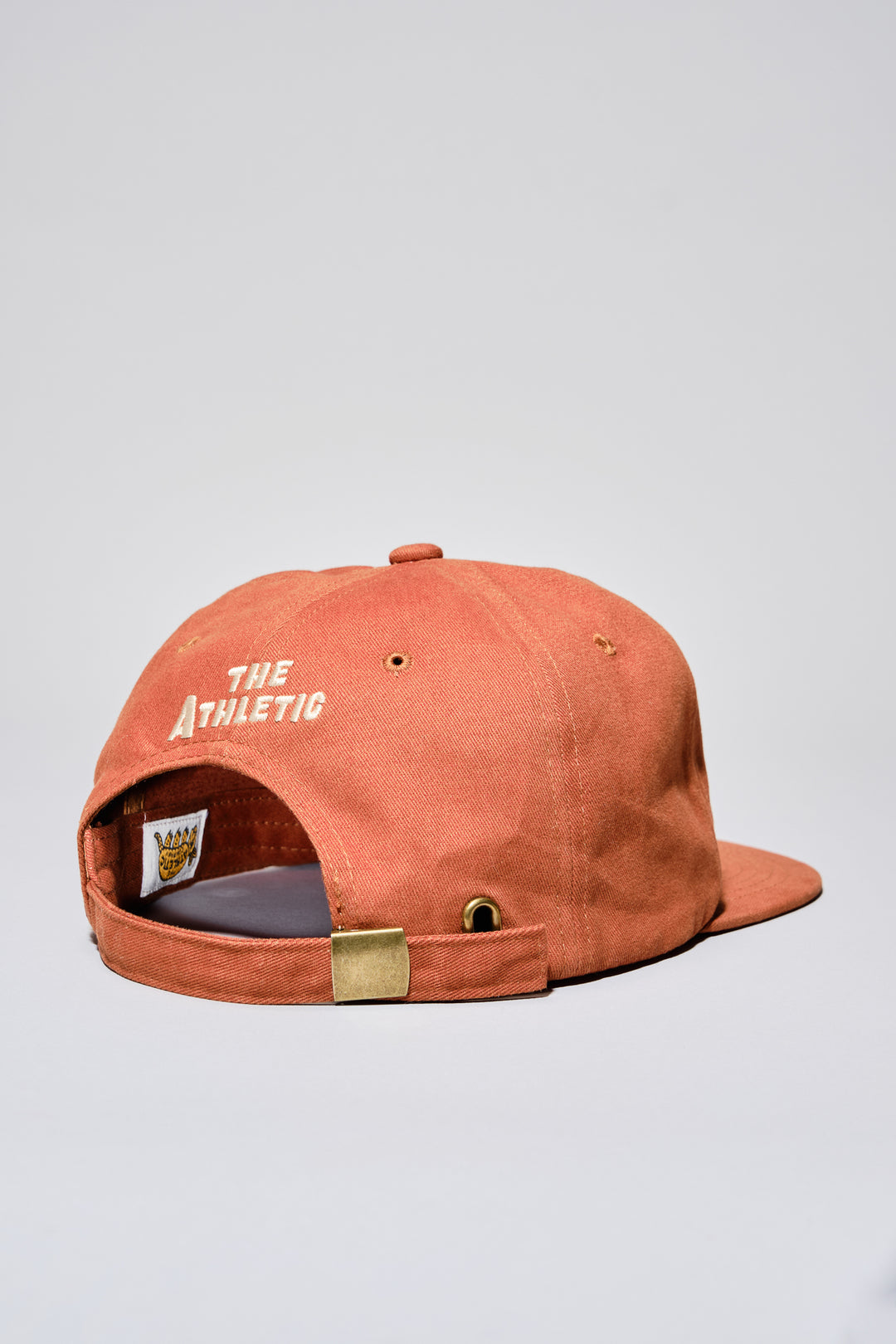 The Athletic Armadillo Logo Hat in rust features an unstructured 6-panel design with an adjustable fabric strap and brass metal clasp. Embroidered with a stylized armadillo logo, this hat combines comfort and style for everyday wear