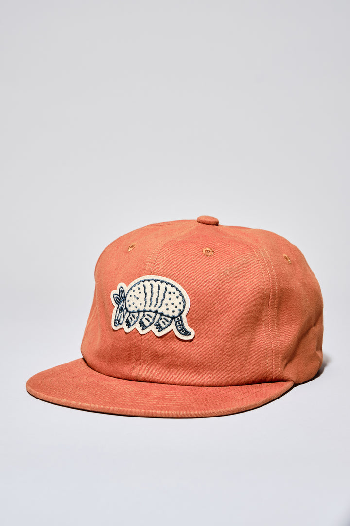 The Athletic Armadillo Logo Hat in rust features an unstructured 6-panel design with an adjustable fabric strap and brass metal clasp. Embroidered with a stylized armadillo logo, this hat combines comfort and style for everyday wear