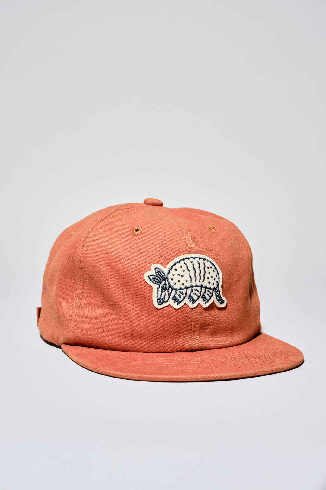 The Athletic Armadillo Logo Hat in rust features an unstructured 6-panel design with an adjustable fabric strap and brass metal clasp. Embroidered with a stylized armadillo logo, this hat combines comfort and style for everyday wear